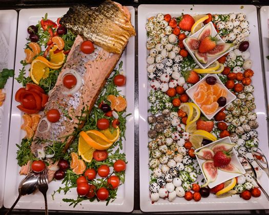 A lavish buffet featuring grilled salmon, fresh sides such as tomatoes and citrus fruits. On the other side, there are attractively decorated platters with fruits and delicious small bites.