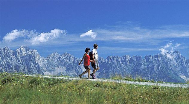Two hikers are walking along a path through a green landscape. In the background, majestic mountains can be seen under a clear blue sky.