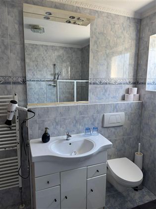 A modern bathroom with a white sink line and a large mirror. The walls are tiled in light colors, and there is a shower as well as hygiene products.