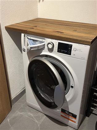 Eine moderne Waschmaschine mit einer Holzoberfläche. Sie steht in einem hellen Raum auf grauen Fliesen.