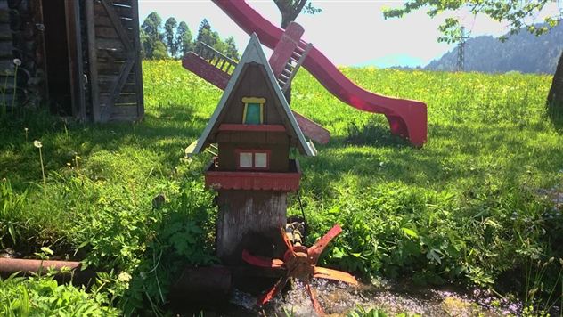 Een kleurrijk speelhuis met een waterwiel staat in een groene tuin. Op de achtergrond zie je een glijbaan en veel bloeiende weilanden.