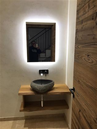 Ein modernes Badezimmer mit einem ovalen Waschbecken aus Stein und einem Holzregal. Der beleuchtete Spiegel und die Holzdetails verleihen dem Raum eine warme Atmosphäre.