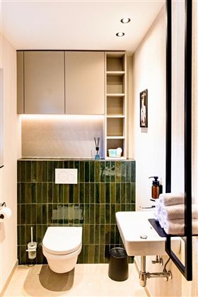 A modern bathroom with green tiles, an elegant toilet, and a sink. The walls are light, and there are practical shelves for storage.