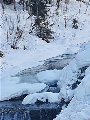 A narrow river flows through a winter landscape with a lot of snow. Some snow-covered rocks lie on the shore.