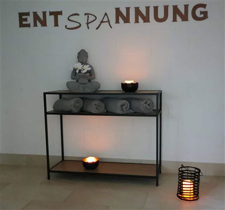 Ein gemütlicher Raum mit einer Buddha-Statue, Handtüchern und Kerzen auf einem Regal. Die Wand hat die Aufschrift „ENTSPANNUNG“ in großen Buchstaben.