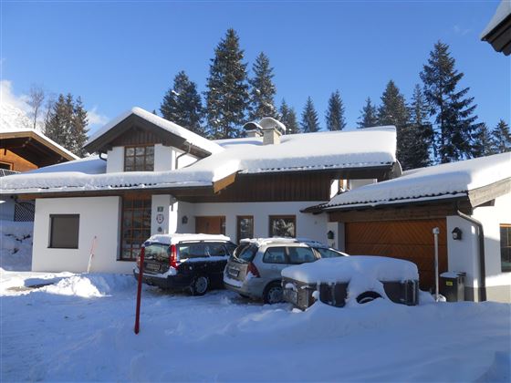 Een modern huis in een sneeuwlandschap met hoge sneeuw. Op de achtergrond zijn naaldbomen en een blauwe lucht te zien.