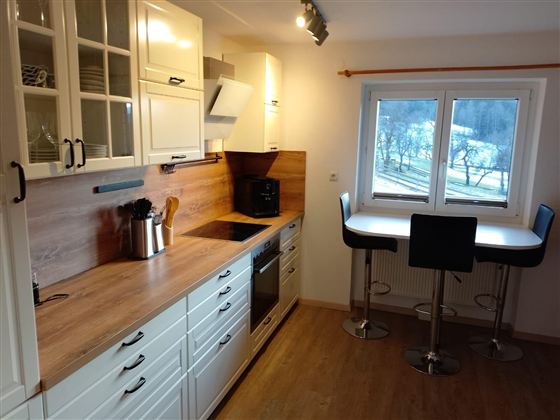 Eine moderne Küche mit Holzoberflächen und weißen Schränken. Sie enthält einen Esstisch mit hohen Stühlen und große Fenster, die Tageslicht hereinlassen.