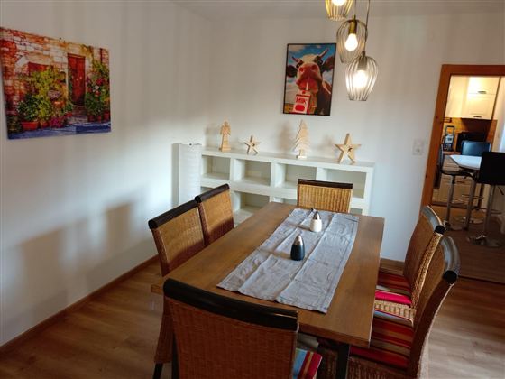 Ein einladender Essbereich mit einem Holztisch und geflochtenen Stühlen. An der Wand hängen zwei bunte Bilder, während der Tisch mit einer Tischdecke und einem Dekorationsobjekt geschmückt ist.