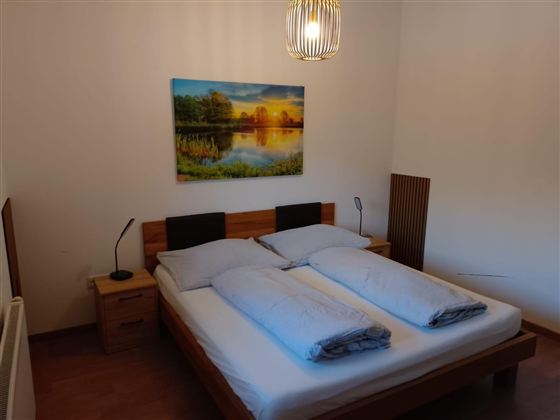 Ein gemütliches Schlafzimmer mit einem Doppelbett und zwei Kissen. An der Wand hängt ein Bild von einer Landschaft mit einem Sonnenuntergang.