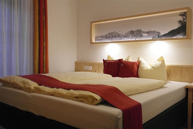 Een gezellige slaapkamer met een groot bed en kleurrijke kussens. Aan de muur hangt een decoratief schilderij, terwijl het licht de ruimte zachtjes verlicht.