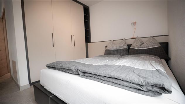 Ein gemütliches Schlafzimmer mit einem modernen Bett und grauer Bettwäsche. Der Schrank an der Wand bietet zusätzlichen Stauraum.