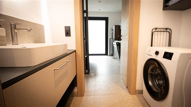 Ein modernes Badezimmer mit einem Waschbecken und einer Waschmaschine. Der Raum hat helle Fliesen und viel natürliches Licht.