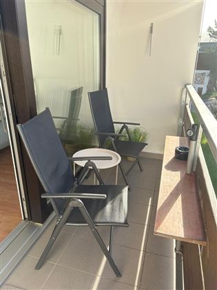 Een ontspannen balkon met twee opvouwbare stoelen en een kleine tafel. De zon schijnt en er staat een groene plant op de achtergrond.