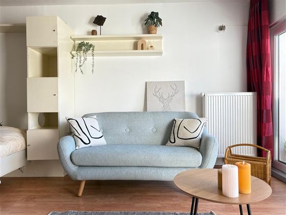 Een moderne woonkamer met een blauwe bank en decoratieve kussens. Een ronde tafel met kaarsen en een lichte wandafwerking zorgt voor een uitnodigende sfeer.