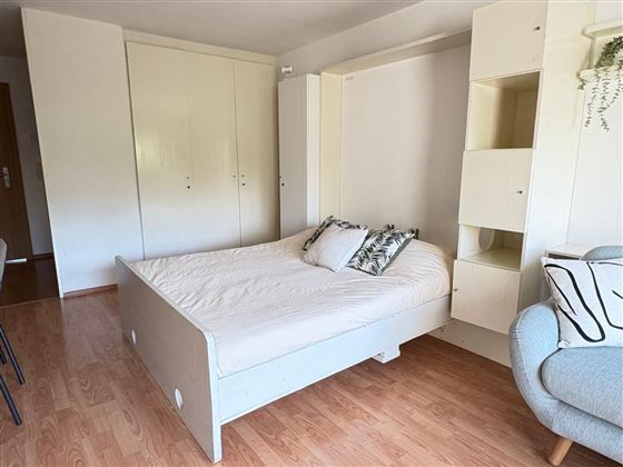 Een lichte kamer met een bed dat goed georganiseerd is. Aan de muur staat een kast en er is een comfortabele stoel in de hoek.