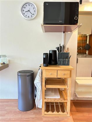 Een compacte keukenopstelling met een houten tafel, magnetron en verschillende keukentoestellen. Naast de tafel staat een prullenbak en een muur met een klok.