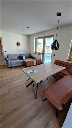 Ein modernes Wohnzimmer mit einem Esstisch und bequemen Sitzgelegenheiten. Große Fenster sorgen für viel Tageslicht und eine einladende Atmosphäre.