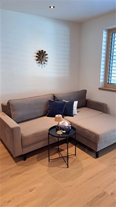 Ein gemütliches Wohnzimmer mit einer grauen Couch und einem kleinen Tisch. Auf der Couch liegen Kissen und ein dekorativer Lampenständer auf dem Tisch.