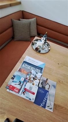 Ein Holztisch mit verschiedenen Broschüren über Kitzbühel und ein Museum. Im Hintergrund sieht man ein gemütliches Sofa und eine dekorative Figur.