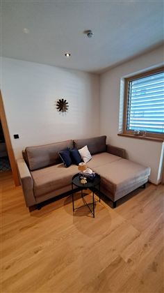 Ein gemütliches Wohnzimmer mit einem modernen Ecksofa und einem kleinen Beistelltisch. Die Fenster lassen viel Licht herein und die Holzfußboden verleiht Wärme.