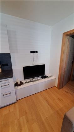 Ein Wohnzimmer mit einem schwarzen Fernseher auf einem weißen TV-Möbel. Der Fußboden ist aus Holz und eine Tür führt zu einem anderen Raum.