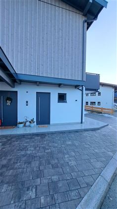 Ein modernes Gebäude mit einer hellen Fassade und grauen Türen. Der gepflasterte Weg vor dem Eingang verleiht dem Bereich ein einladendes Aussehen.