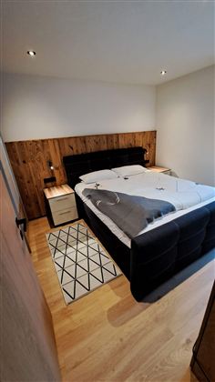Ein modernes Schlafzimmer mit einem großen Bett und einer Holzwand. Der Raum hat einen warmen Holzfußboden und ein dekoratives Teppichmuster.