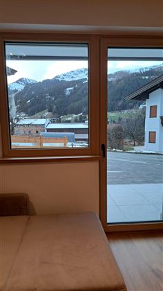 Ein Fenster mit Blick auf schneebedeckte Berge und eine ruhige Landschaft. Im Vordergrund ist ein gemütliches Sofa zu sehen.