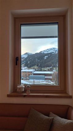 Ein Fenster mit Blick auf schneebedeckte Berge und ein typisches alpenländisches Dorf. Der Himmel ist leicht bewölkt und die Umgebung wirkt ruhig.