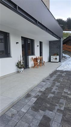 Eine moderne Terrasse mit einem kleinen Tisch und zwei Stühlen. Der Boden ist aus grauen Steinen, und es liegt noch etwas Schnee.