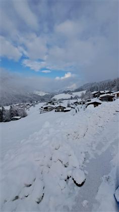 Eine verschneite Landschaft mit Hütten und Bäumen. Der Himmel ist teilweise sonnig und wolkig.