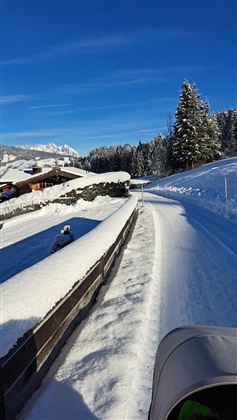 Eine verschneite Straße in einer malerischen Landschaft. Im Hintergrund sind schneebedeckte Bäume und Hütten zu sehen.