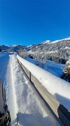 Eine verschneite Landschaft mit Bergen im Hintergrund und klarem blauem Himmel. Der Weg ist von frischem Schnee bedeckt.
