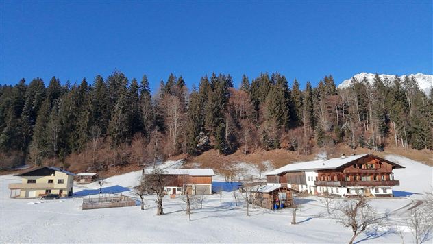 Een winterlandschap met verschillende hutten en met sneeuw bedekte gebieden. Op de achtergrond rijzen hoge, beboste bergen op onder een stralend blauwe lucht.