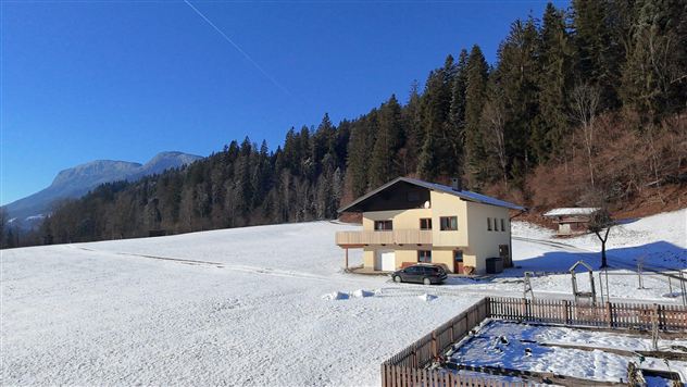 Een gezellig huis staat op een met sneeuw bedekt veld, omgeven door bomen. Op de achtergrond zijn bergen en een heldere blauwe lucht zichtbaar.