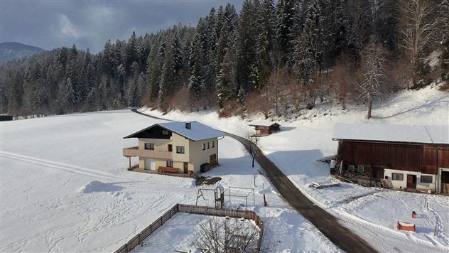 Een besneeuwd landschap met een woonhuis en aangrenzende gebouwen. Op de achtergrond zijn hoge bomen en een heldere lucht te zien.