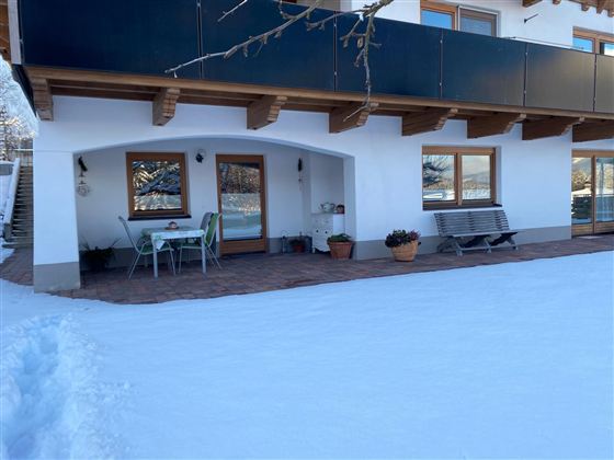 Ein gemütlicher Patio an einem Haus, umgeben von Schnee. Tische und Stühle laden zum Entspannen ein.
