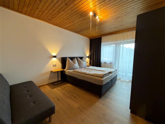 Een sfeervolle slaapkamer met een tweepersoonsbed en twee kussens. Er is een bank en grote ramen die veel daglicht binnenlaten.