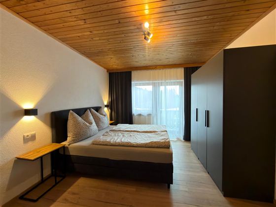 Een moderne slaapkamer met een groot bed en houten plafond. De ruimte heeft grote ramen en een stijlvolle kast.