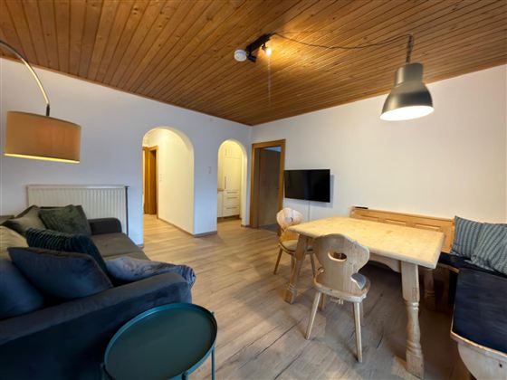 Een gezellige woonkamer met een bank, een tafel en stoelen. De kamer heeft een lichte houten plafond en een televisie aan de muur.