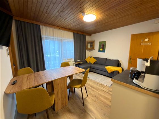 Een gezellige woonkamer met een eettafel en gele stoelen. Op de achtergrond bevindt zich een bank en een raamkant die natuurlijk licht binnenlaat.