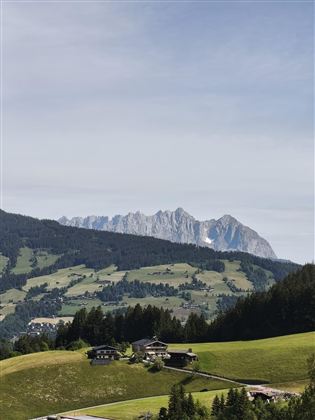 Eine idyllische Berglandschaft mit sanften Hügeln und wenigen Gebäuden im Vordergrund. Im Hintergrund sind beeindruckende Felsformationen zu sehen.