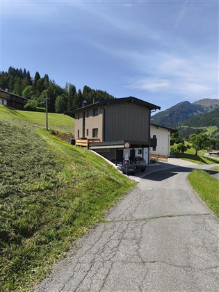 Ein modernes Haus steht an einer kurvenreichen Straße in einer grünen Landschaft. Im Hintergrund sind sanfte Hügel und Bäume sichtbar.