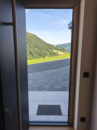 Eingangstür mit Blick auf eine grüne Hügel-Landschaft. Der Himmel ist blau und die Straße führt ins Tal.