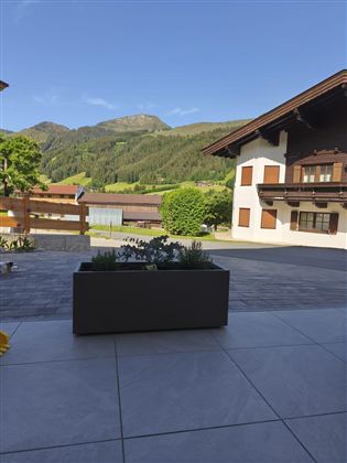 Eine gemütliche Terrasse mit einem Pflanzkübel und Blick auf grüne Hügel. Im Hintergrund sind traditionelle Häuser und ein klarer blauer Himmel zu sehen.
