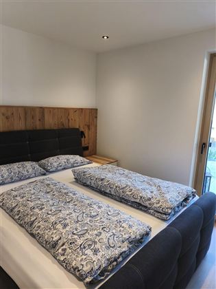 Ein gemütliches Schlafzimmer mit einem Doppelbett und blauen Bettdecken. Der Raum hat einen modernen Look und viel Tageslicht durch ein Fenster.