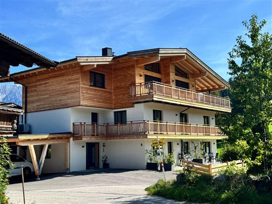 A modern wooden house with several balconies and green surroundings. The sun is shining under a blue sky on the building.