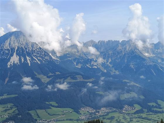 Eine beeindruckende Berglandschaft mit hohen Gipfeln und weißen Wolken. Im Vordergrund sind grüne Wiesen und kleine Siedlungen zu sehen.