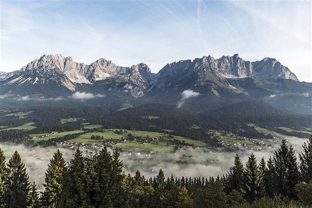 Eine beeindruckende Berglandschaft mit hohen Gipfeln und einem klaren Himmel. Im Vordergrund sind Wälder und eine grüne Hügellandschaft zu sehen.