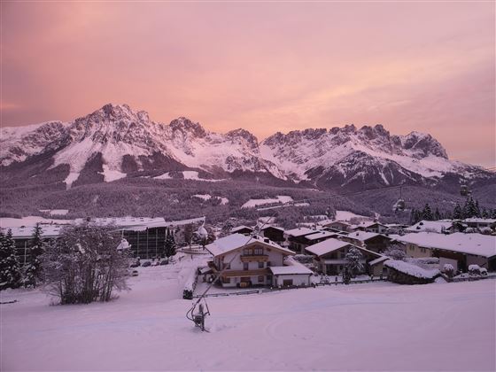 A picturesque winter landscape with snow-covered mountains and a pink sky. In the foreground, cozy houses can be seen in a snowy environment.